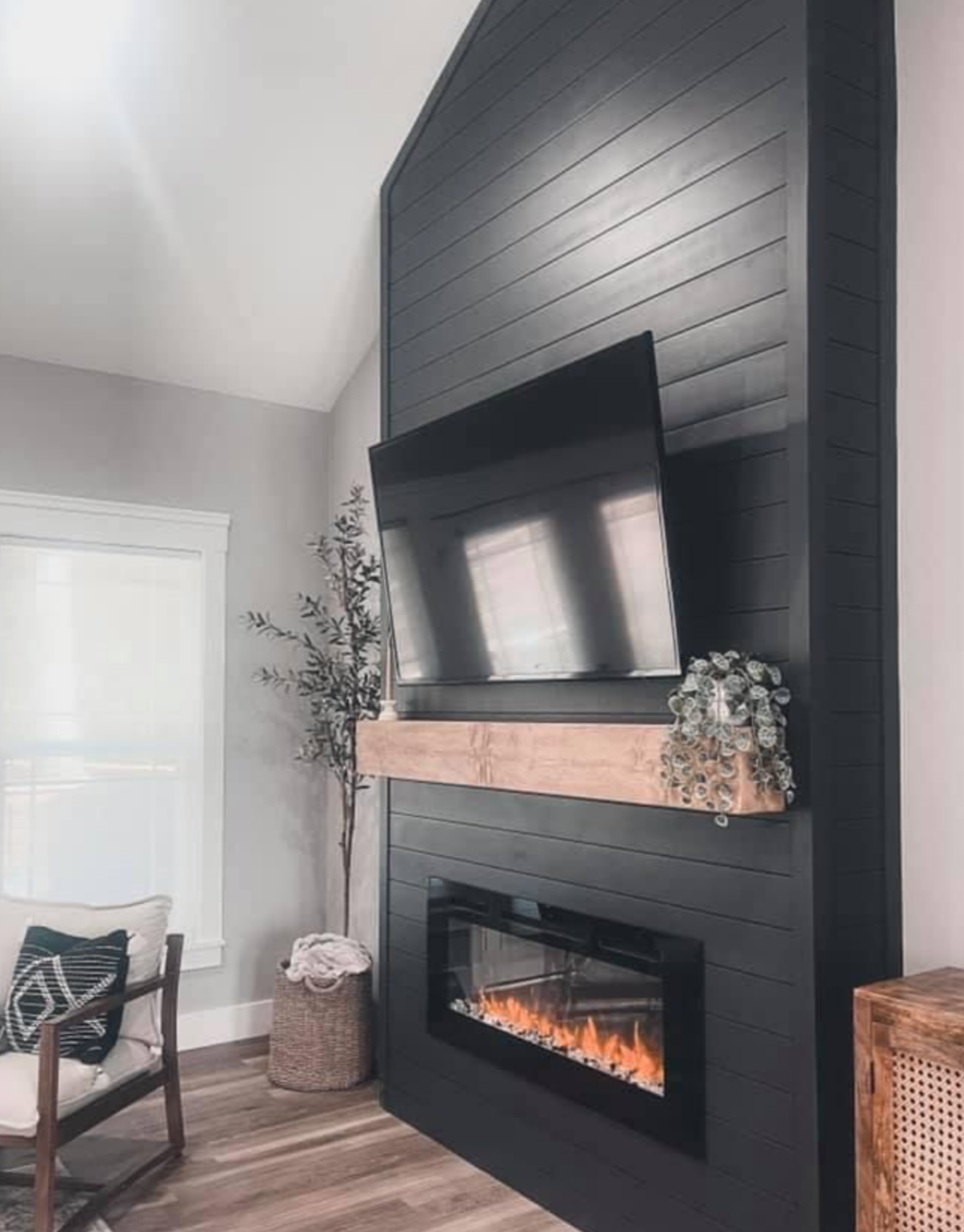 Living room corner with a mounted flat-screen TV on a black wood-paneled wall, a fireplace below, a wooden mantel with decorative flowers, a potted plant, a window with white trim, and a chair with a pillow.