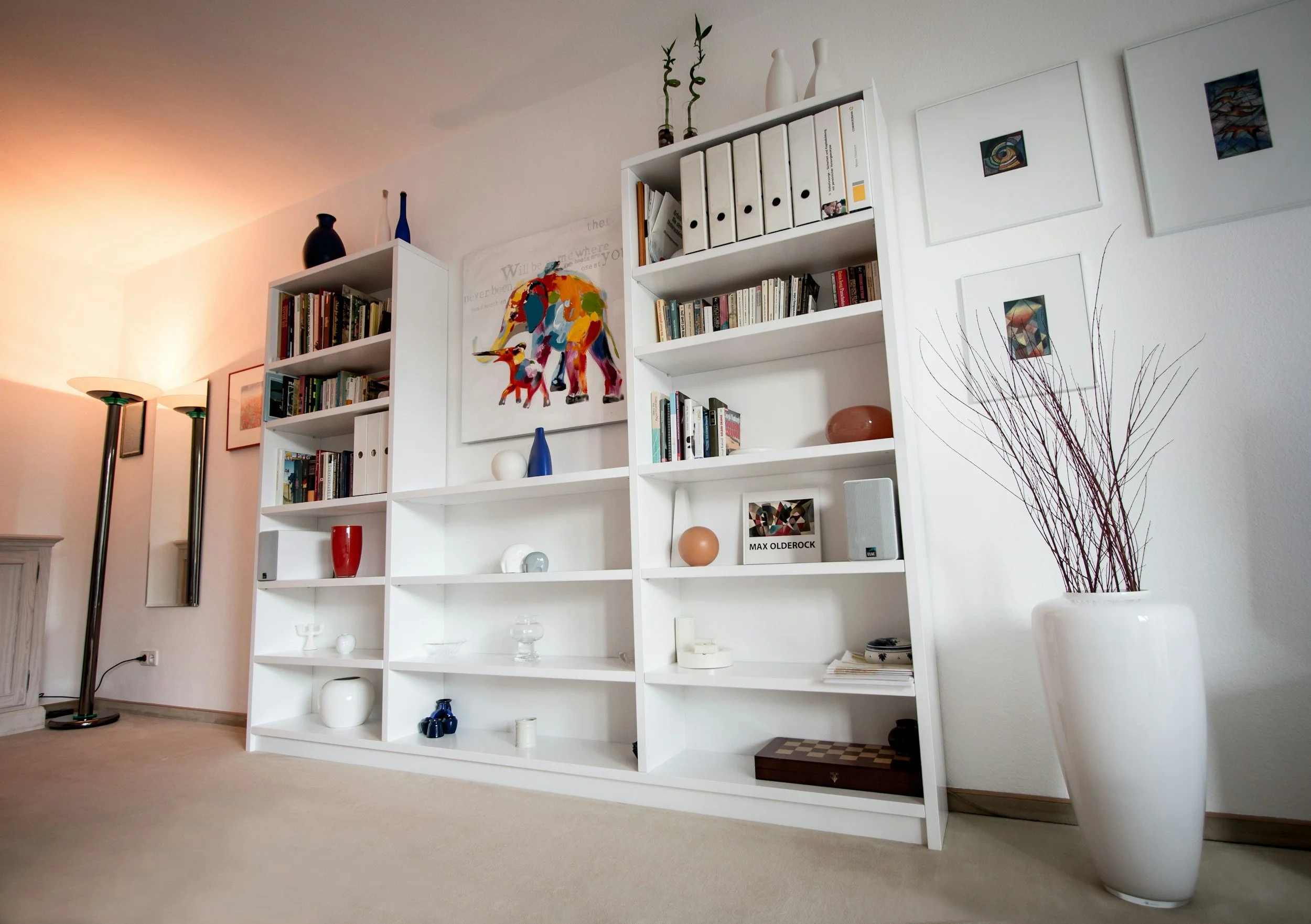 Living room corner with a white shelving unit holding books and decorative objects, a white vase with branches on the floor, a floor lamp with two round lights, and framed artwork on the wall.