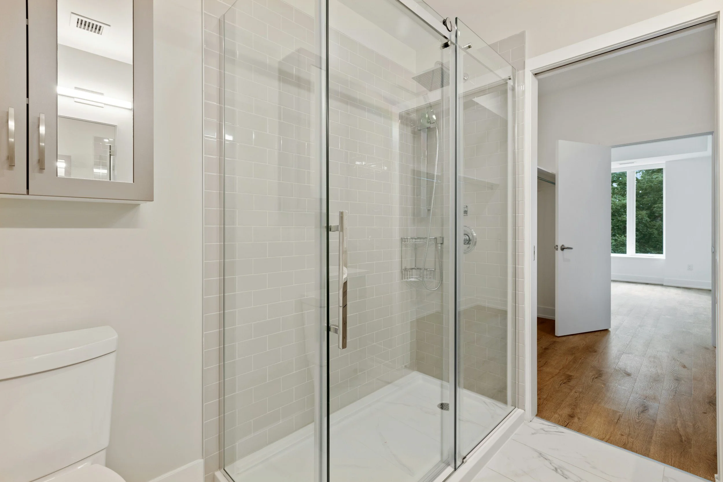 Bathroom with a glass-enclosed shower, a toilet, white tile walls, wooden flooring in the adjacent room, and a large window in the next room.