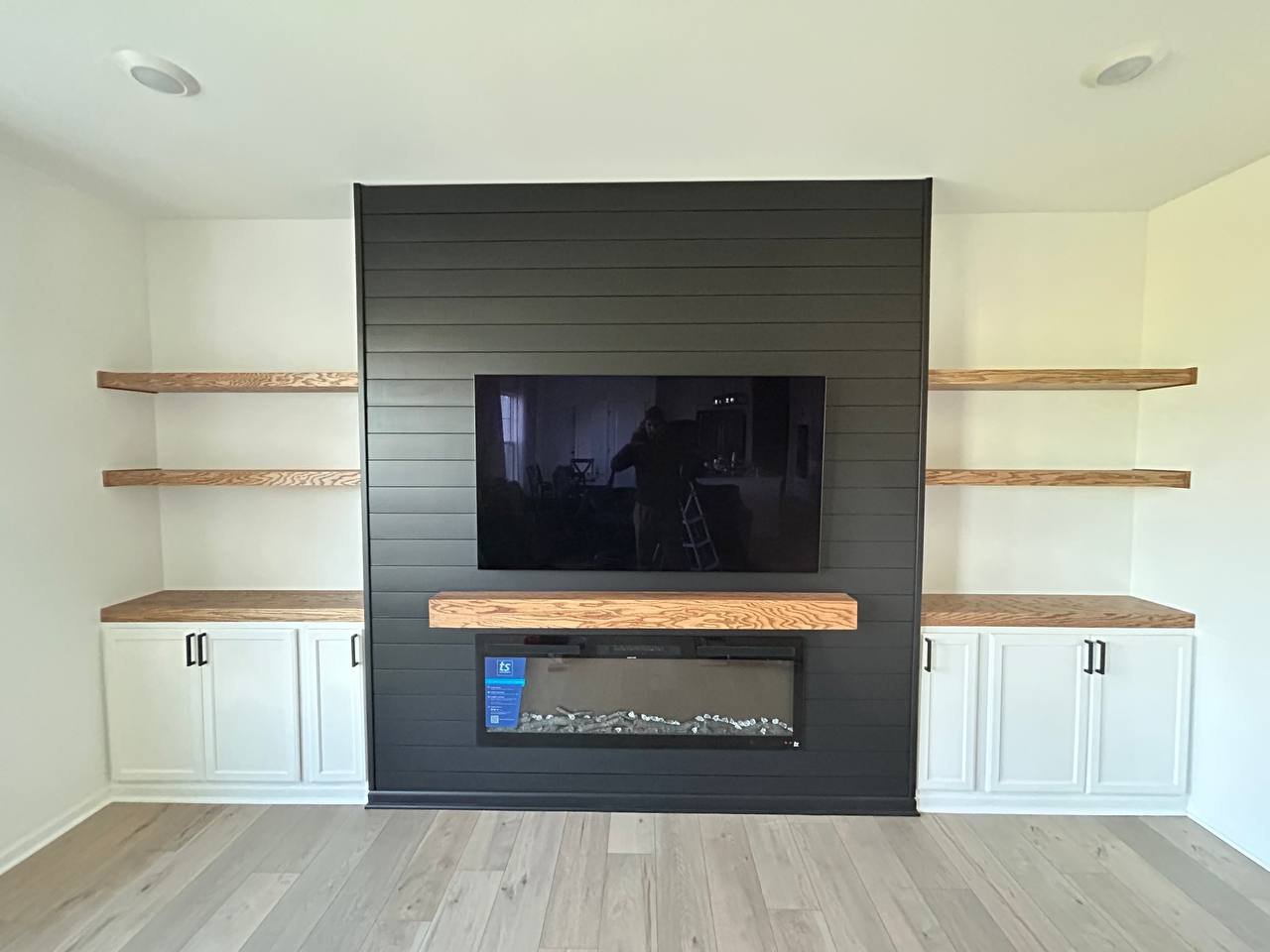 Living room with a black accent wall featuring a mounted flat-screen TV and a modern fireplace beneath it. Light wood built-in shelves on either side of the accent wall. Light-colored hardwood flooring.