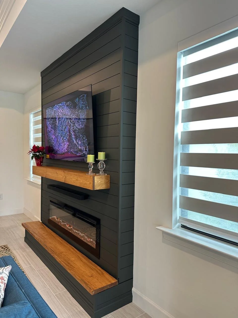 Living room with a flat-screen TV mounted on a dark gray shiplap wood wall, a wooden mantel with candles, a fireplace below with decorative elements, and a window with zebra blinds.