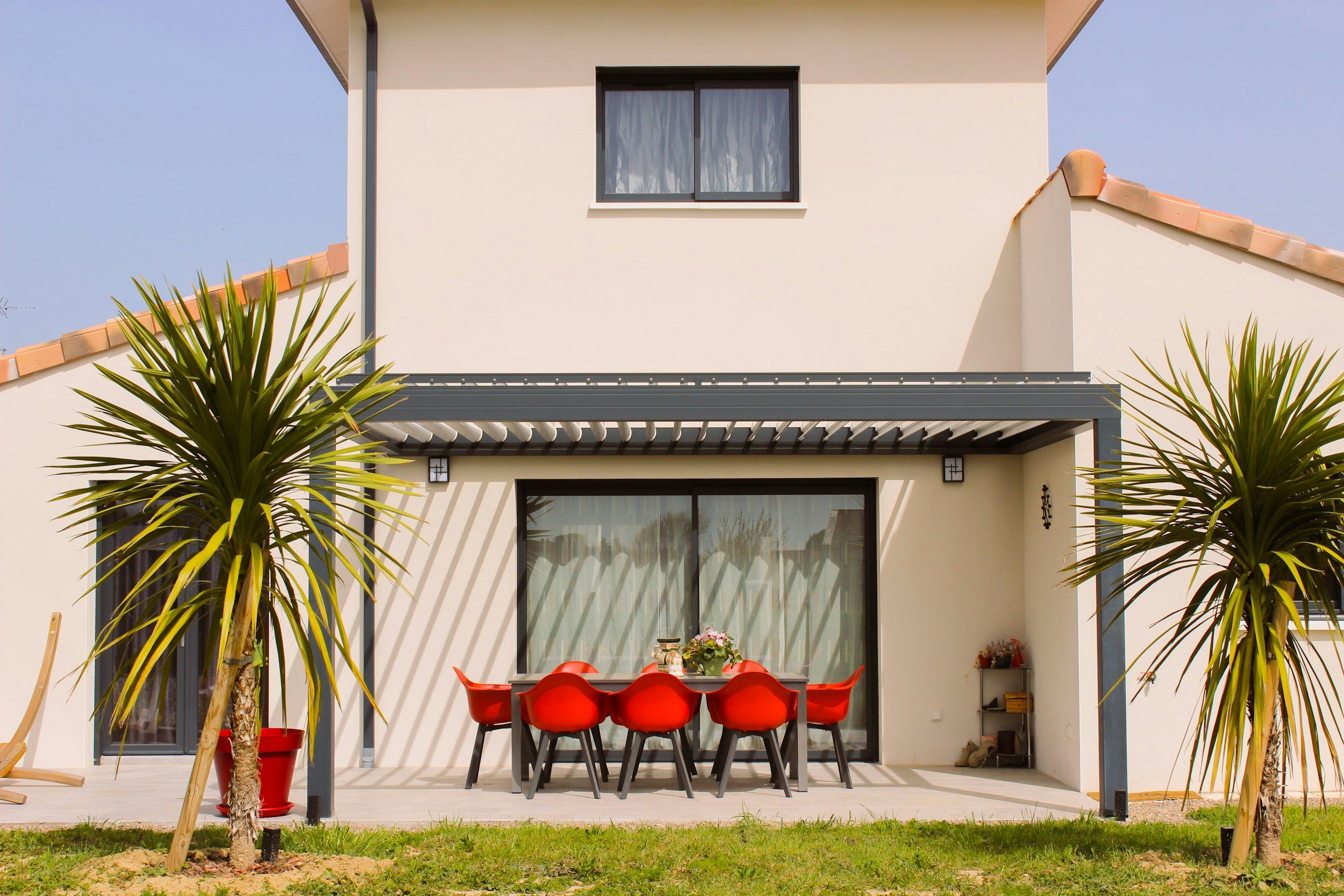 Exterior view of a modern two-story house with a patio. The house has white walls, a large sliding glass door, and a window on the upper floor. The patio features a dining table with eight red chairs, shaded by a pergola. Two palm trees are planted in the yard, and there is a small shelf with books and decorative items on the right side of the patio.