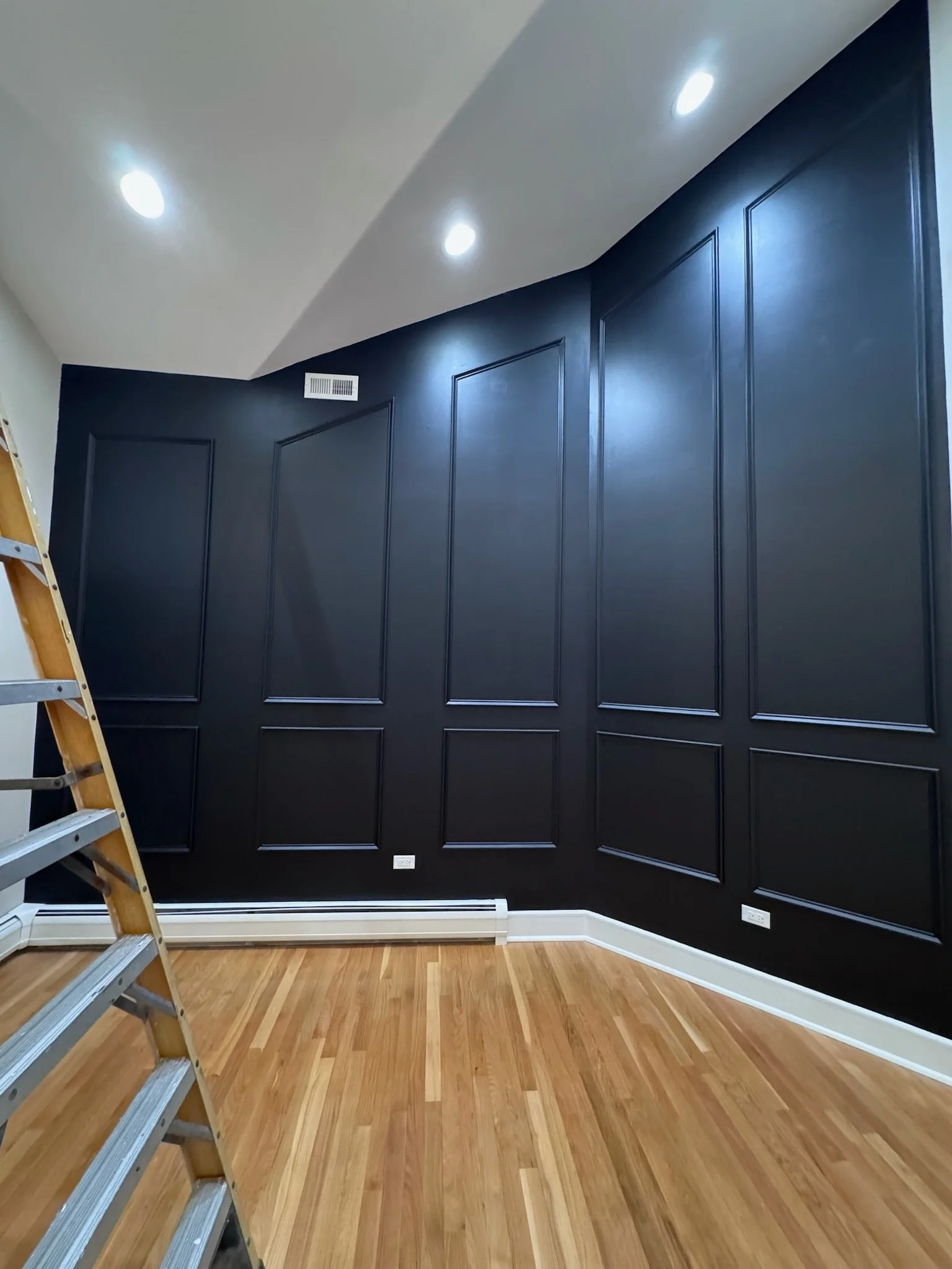 Interior room with dark blue walls featuring decorative molding, hardwood floor, white baseboards, and ceiling with recessed lighting. Part of a wooden ladder is visible on the left side.