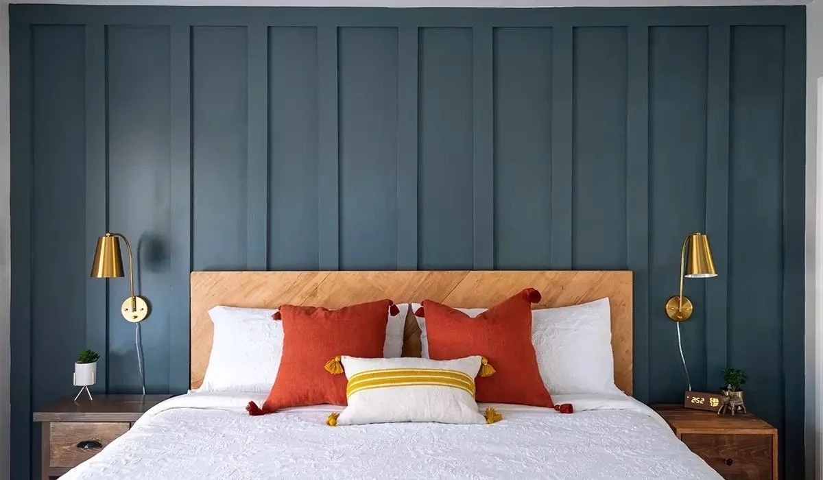 A bedroom with a wooden headboard, blue paneled wall, and white bedding. There are two gold wall-mounted lamps, and decorative pillows in red, orange, and yellow with tassels. Small potted plants are on each side on nightstands.