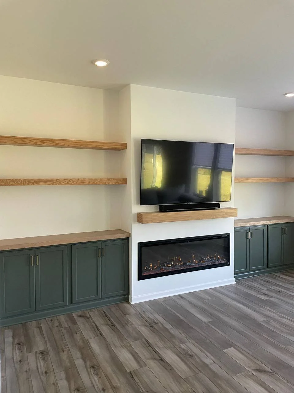 Living room with white walls, hardwood flooring, a mounted flat-screen TV, an electric fireplace beneath it, wooden shelves on both sides, and green cabinetry below the shelves.