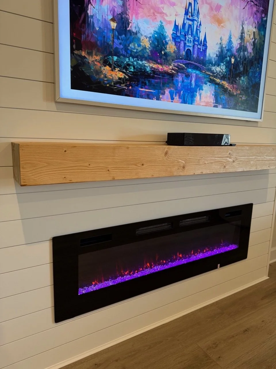 A living room wall with a flat-screen television displaying a colorful digital painting of a castle and landscape, a wooden mantel shelf beneath the TV, and a black electric fireplace with purple and orange flames located below the shelf.