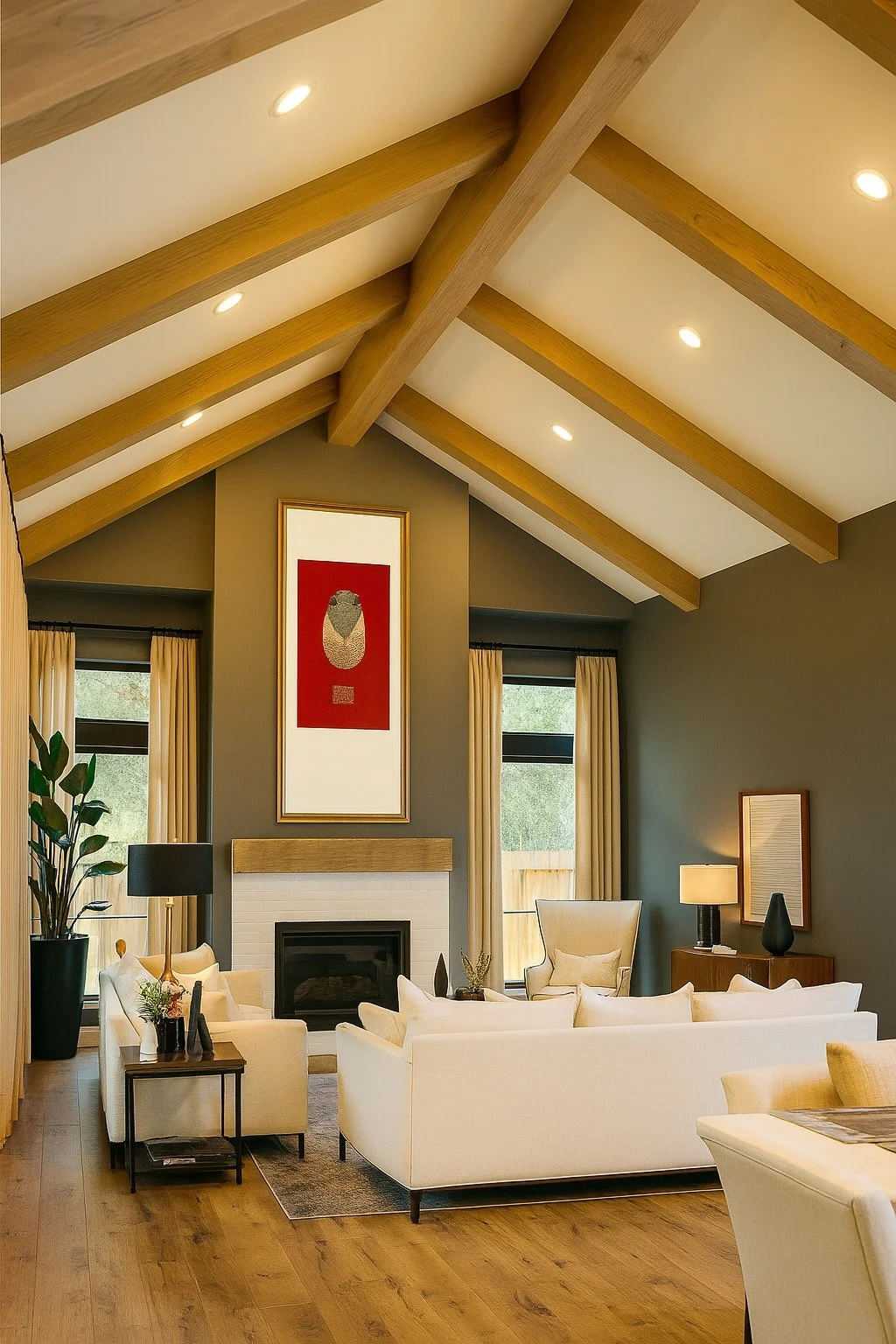 Living room with a vaulted ceiling featuring exposed wooden beams, a fireplace with a wooden mantel, cream-colored sofas, and large windows with cream curtains. Decor includes a tall potted plant, framed artwork above the fireplace, and side tables w