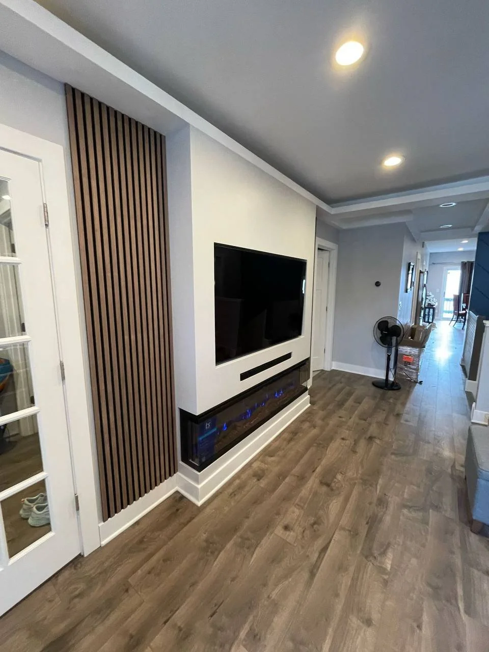 Modern living room with wood flooring, white walls, TV mounted on the wall, electric fireplace below the TV, wooden slat accent wall on the left, ceiling recessed lights, and a standing fan in the background.