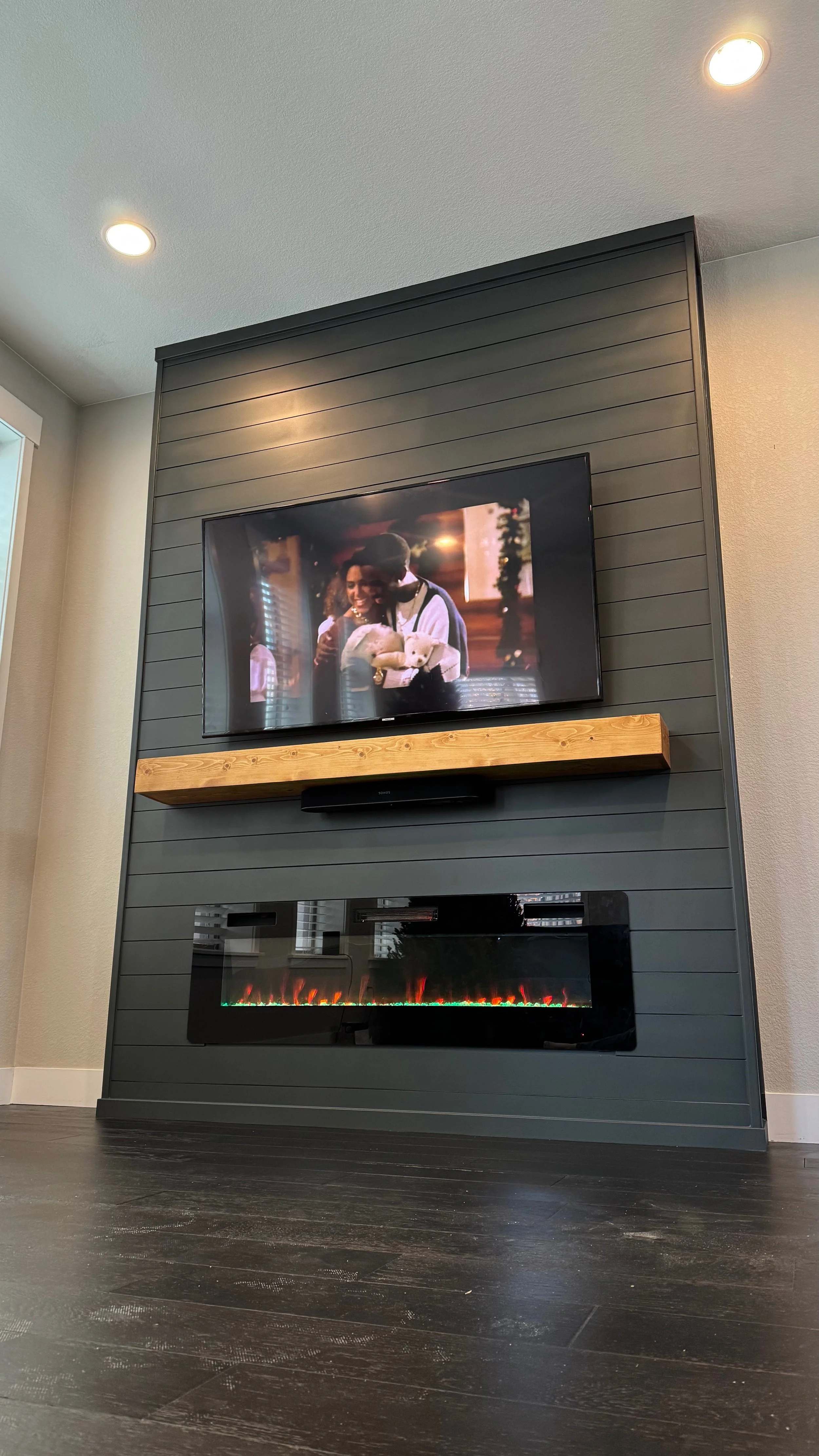 A living room corner with a black entertainment wall featuring a mounted TV showing a scene with women hugging, and a built-in electric fireplace below it, with a wooden shelf in between, black flooring, and ceiling recessed lights.