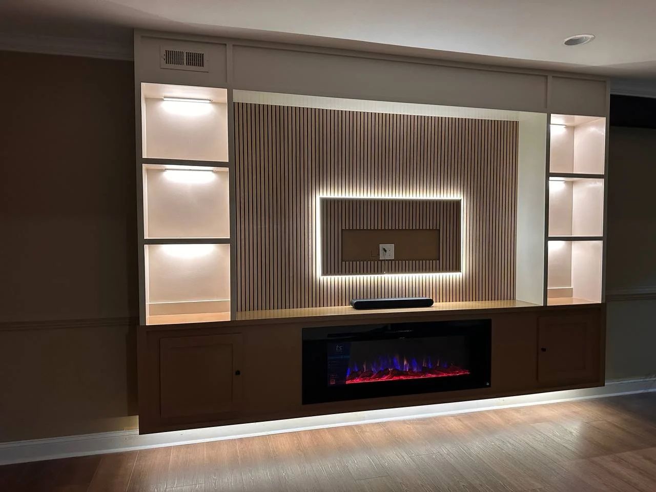 Modern entertainment wall with built-in shelves and an electric fireplace, wood paneling, and backlit LED lighting.
