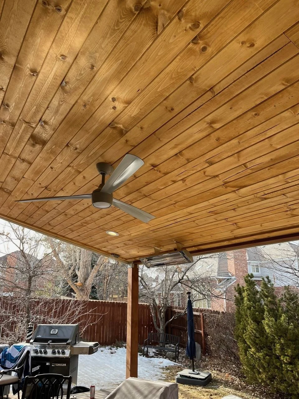 Covered backyard patio with wooden ceiling, ceiling fan, outdoor grill, and outdoor furniture, surrounded by trees and a wooden privacy fence.