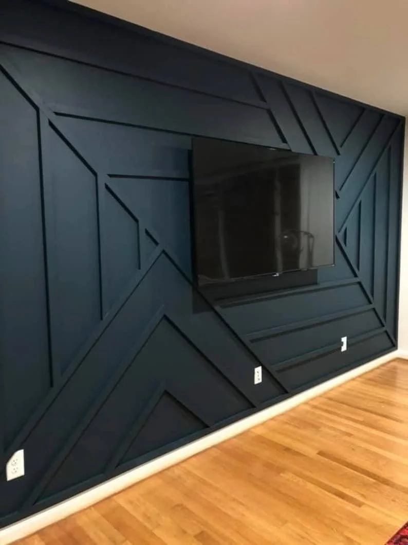 A flat-screen television mounted on a dark geometric wood panel wall in a living room, with hardwood flooring.