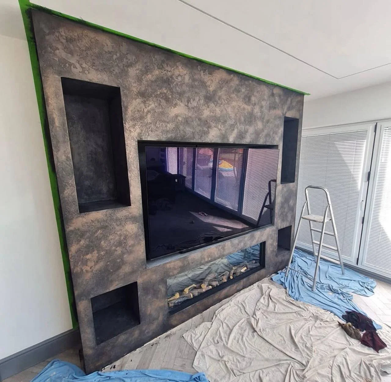 Living room with a modern fireplace and built-in shelves, with painting and construction materials on floor, ladder nearby, and window blinds.