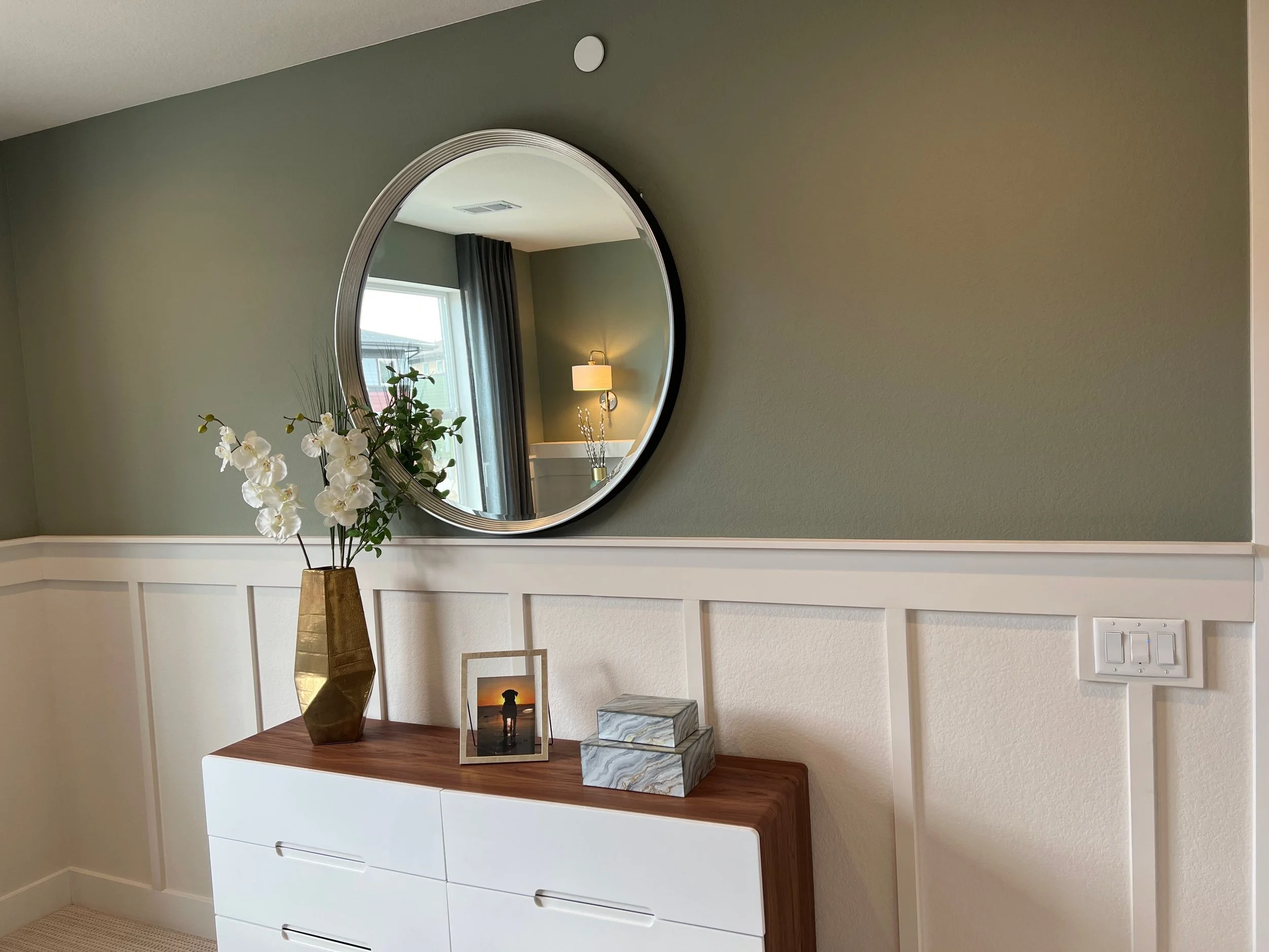 Interior of a modern room featuring a white dresser, a tall gold geometric vase with white artificial orchids, a framed photo of a dog at sunset, and two decorative marble boxes on top. A round mirror hangs on a green wall, reflecting a window with dark curtains, a white lamp, and a wall sconce.