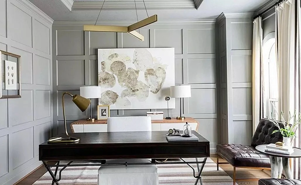 Modern home office with gray paneled walls, a white desk, and a black leather lounge chair near a window with white curtains.