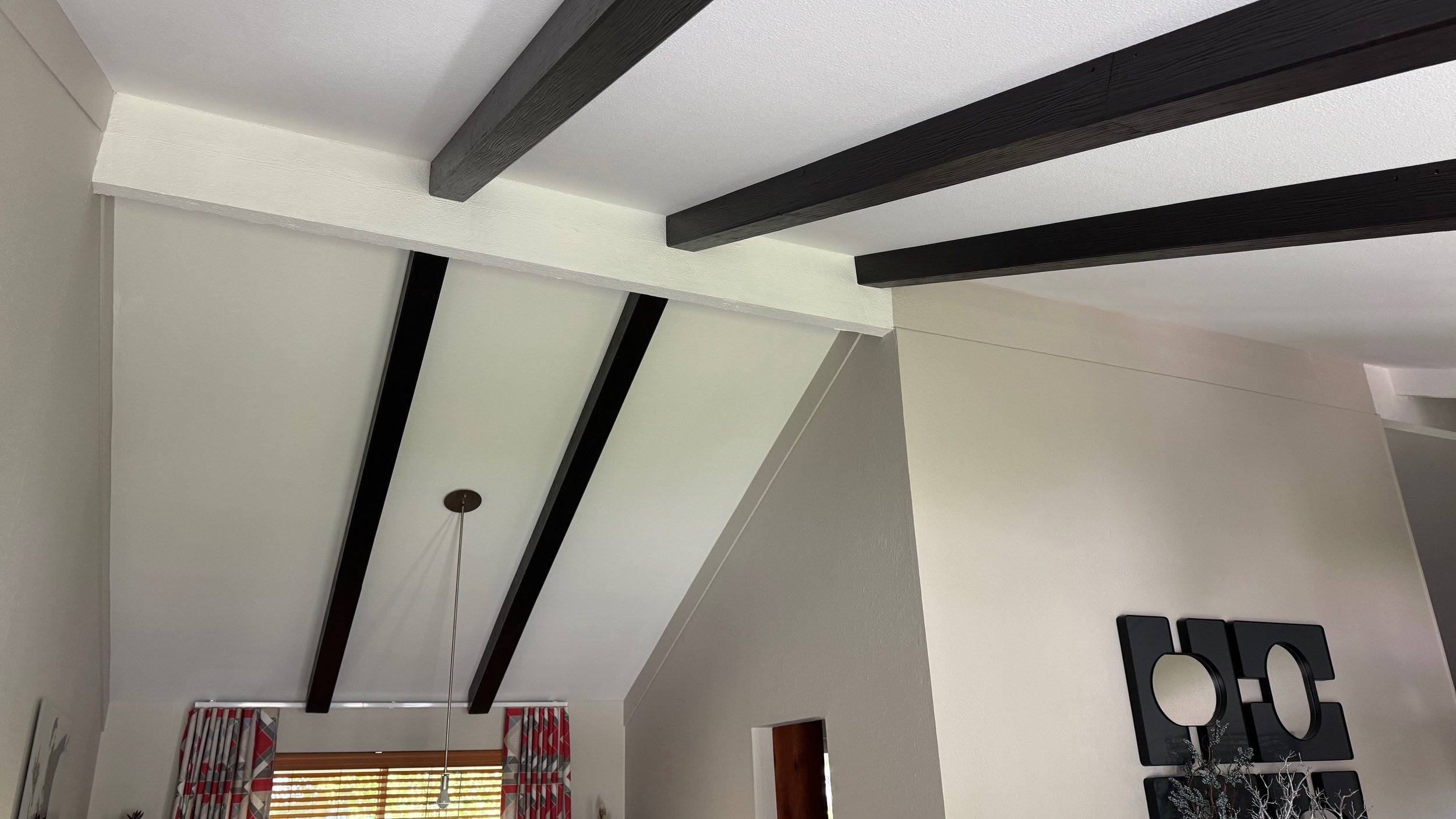 Interior ceiling with exposed dark wooden beams, white ceiling panels, and a hanging light fixture, with a window and curtains in the background.