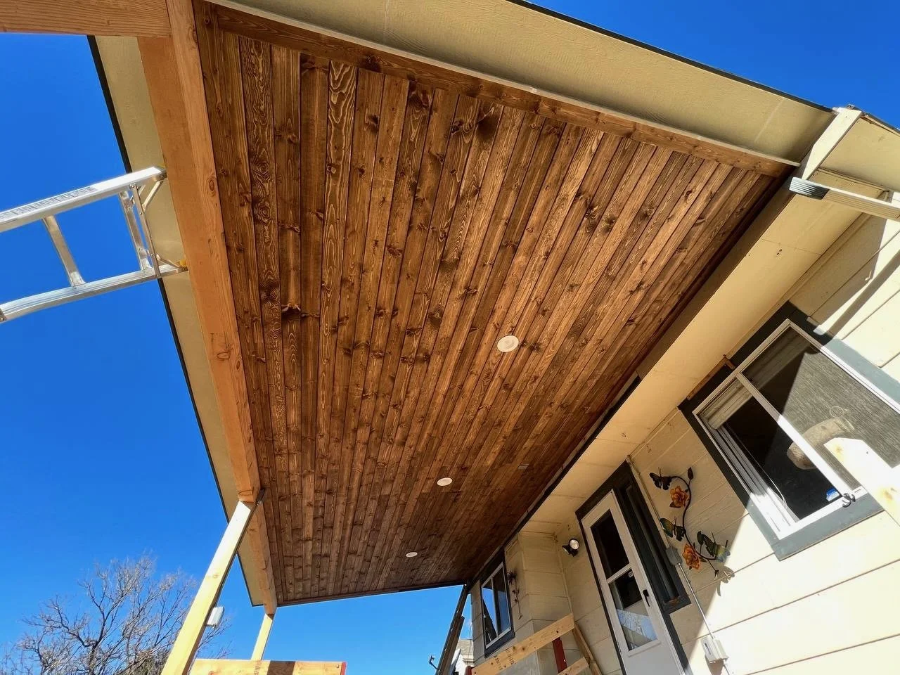 Under construction porch with unfinished wooden ceiling and support beams, ladder leaning against the structure, and house exterior with windows and decorative wall flower ornament.