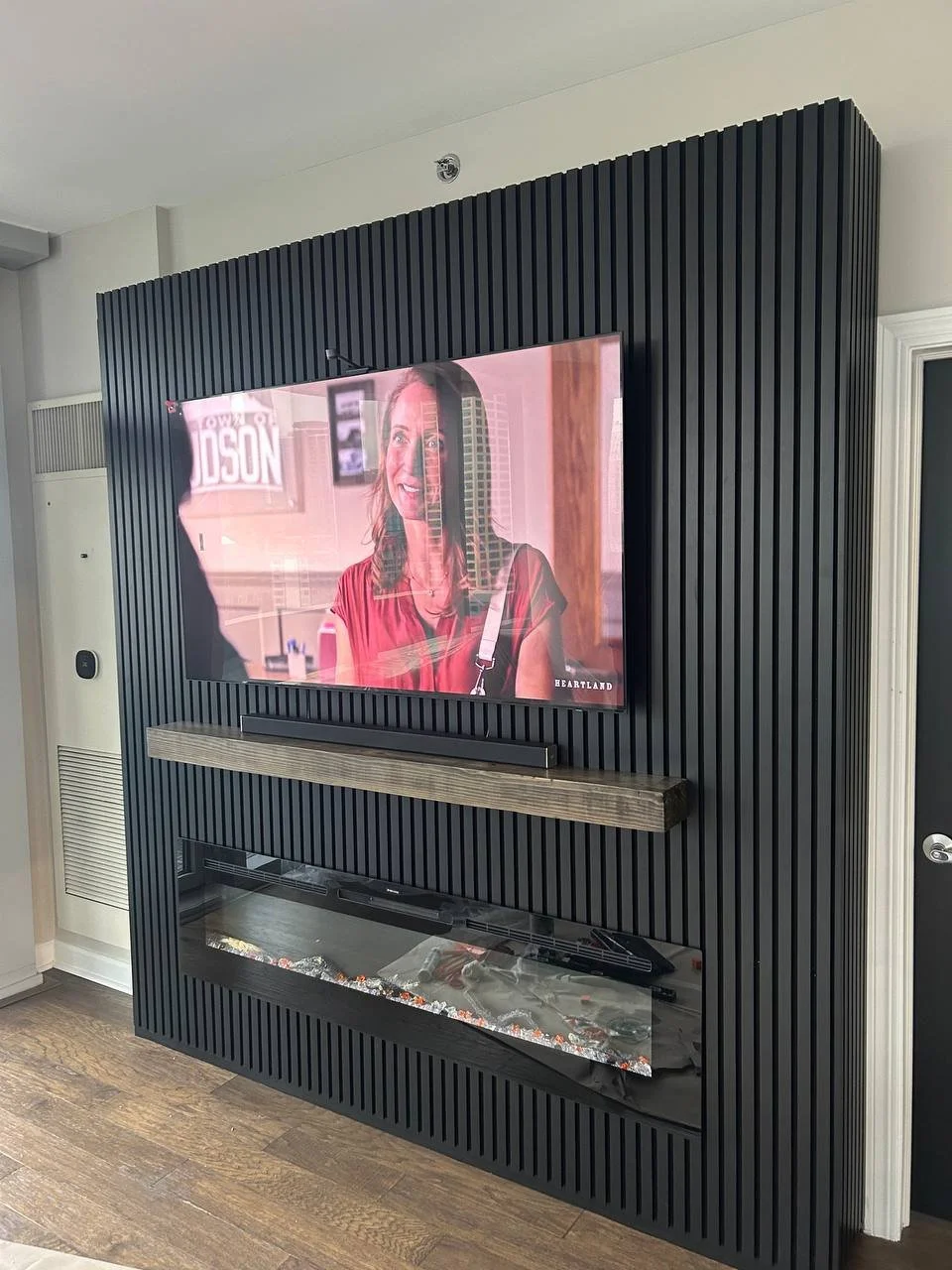 A living room wall with a mounted flat-screen television displaying a woman in a red dress, a wooden shelf, and a modern electric fireplace beneath the shelf, all set against a black, vertically paneled wall.