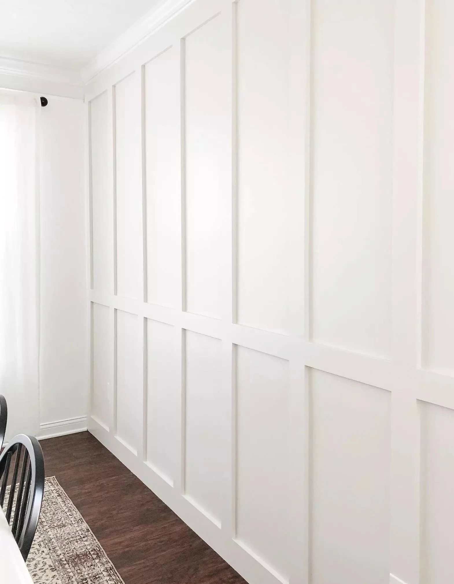 White interior wall with decorative wood paneling, dark hardwood floor, portion of a dining table, and chairs.