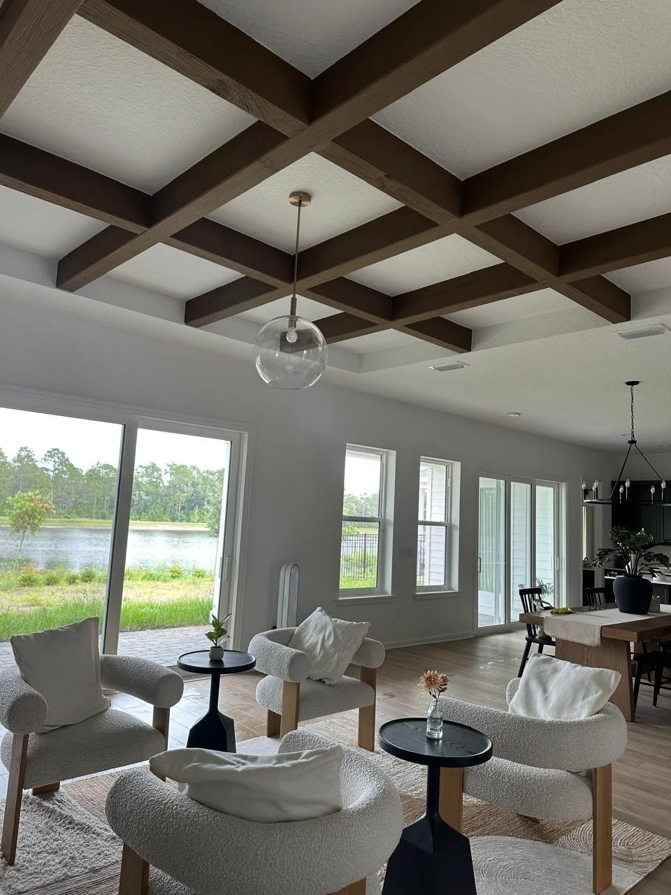 Bright living room with large windows overlooking a lake, featuring white armchairs, small black side tables, a beige area rug, and a modern ceiling grid with wooden beams.