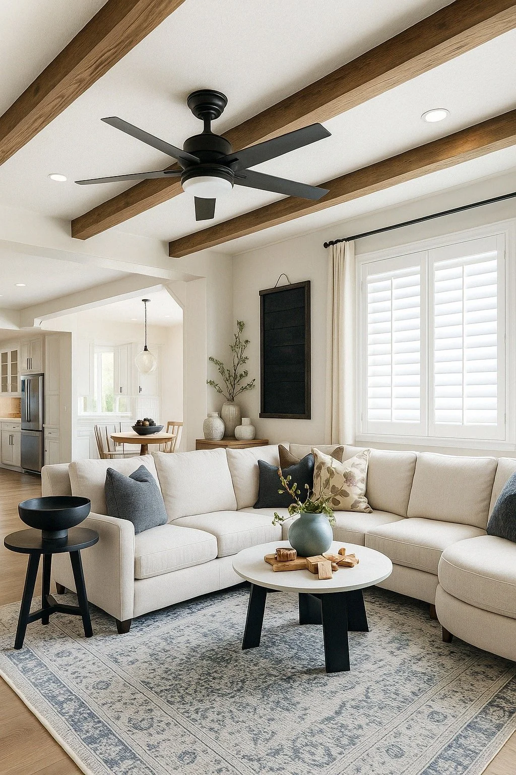 Living room with a white sectional sofa, black side table, coffee table with a vase of flowers, wood ceiling beams, window with white shutters, and a ceiling fan.