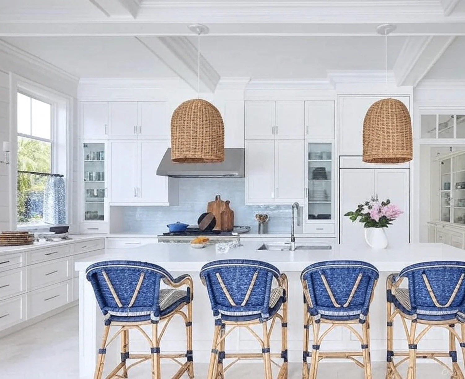 Bright white kitchen with large island, blue rattan chairs, wicker pendant lights, white cabinetry, and a window with view of greenery.