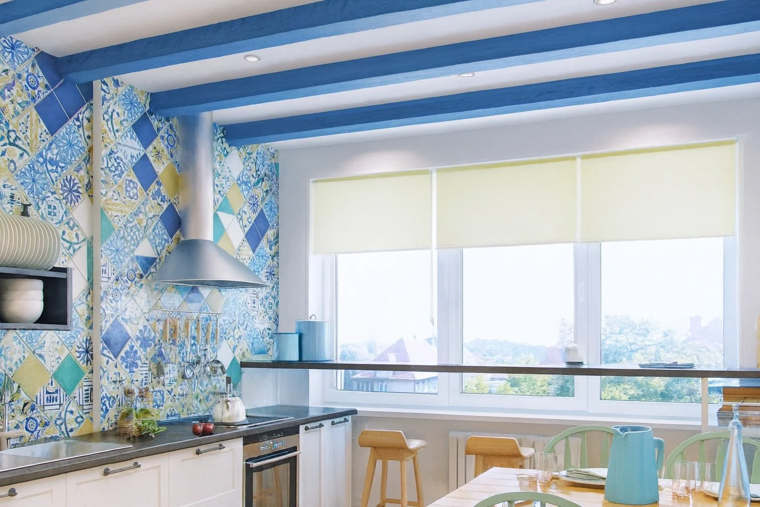 Bright kitchen with blue patterned tiled wall, blue ceiling beams, white cabinets, stove, large window with yellow blinds, dining table with green chairs, and various kitchen accessories.