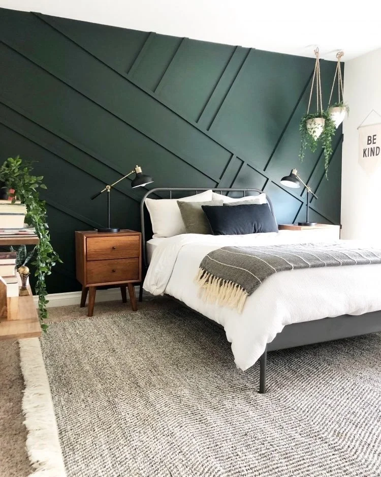 Modern bedroom with dark green geometric accent wall, white bedding, and wooden nightstands with black desk lamps, decorated with hanging plants and framed wall art.