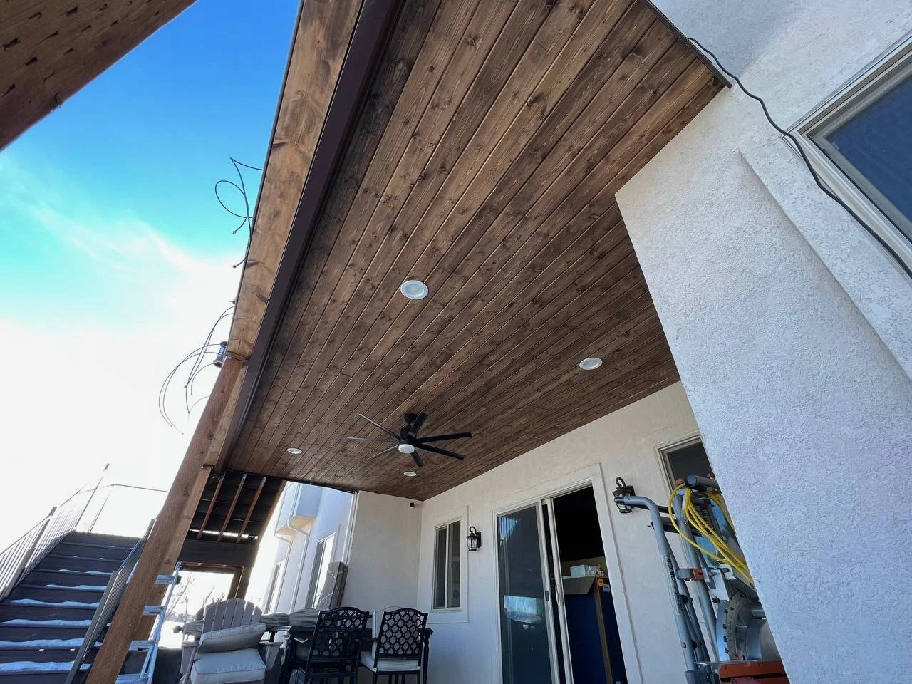 View of a partially covered patio with a wooden ceiling, a ceiling fan, white walls, a window, and an outdoor staircase leading up to a second level, with some construction materials and wiring visible.
