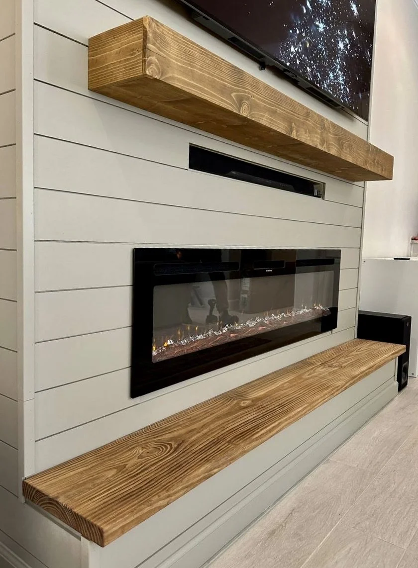 A modern electric fireplace with a black frame built into a white paneled wall, flanked by two long wooden shelves above and below.