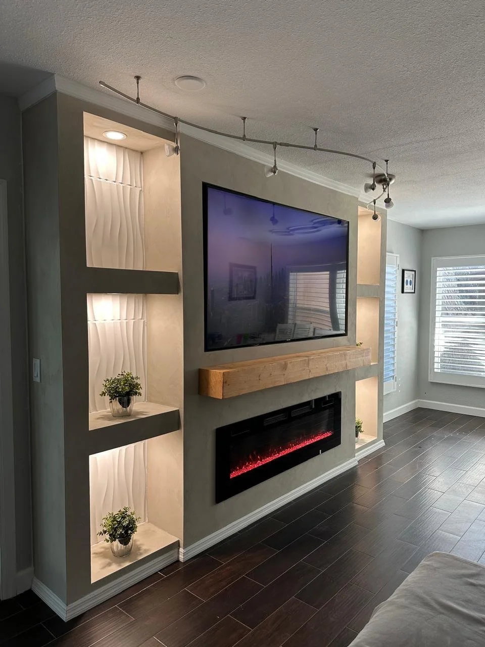 Living room with a wall-mounted TV, a decorative fireplace below it, and built-in shelves with plants on the sides, illuminated with spotlighting.