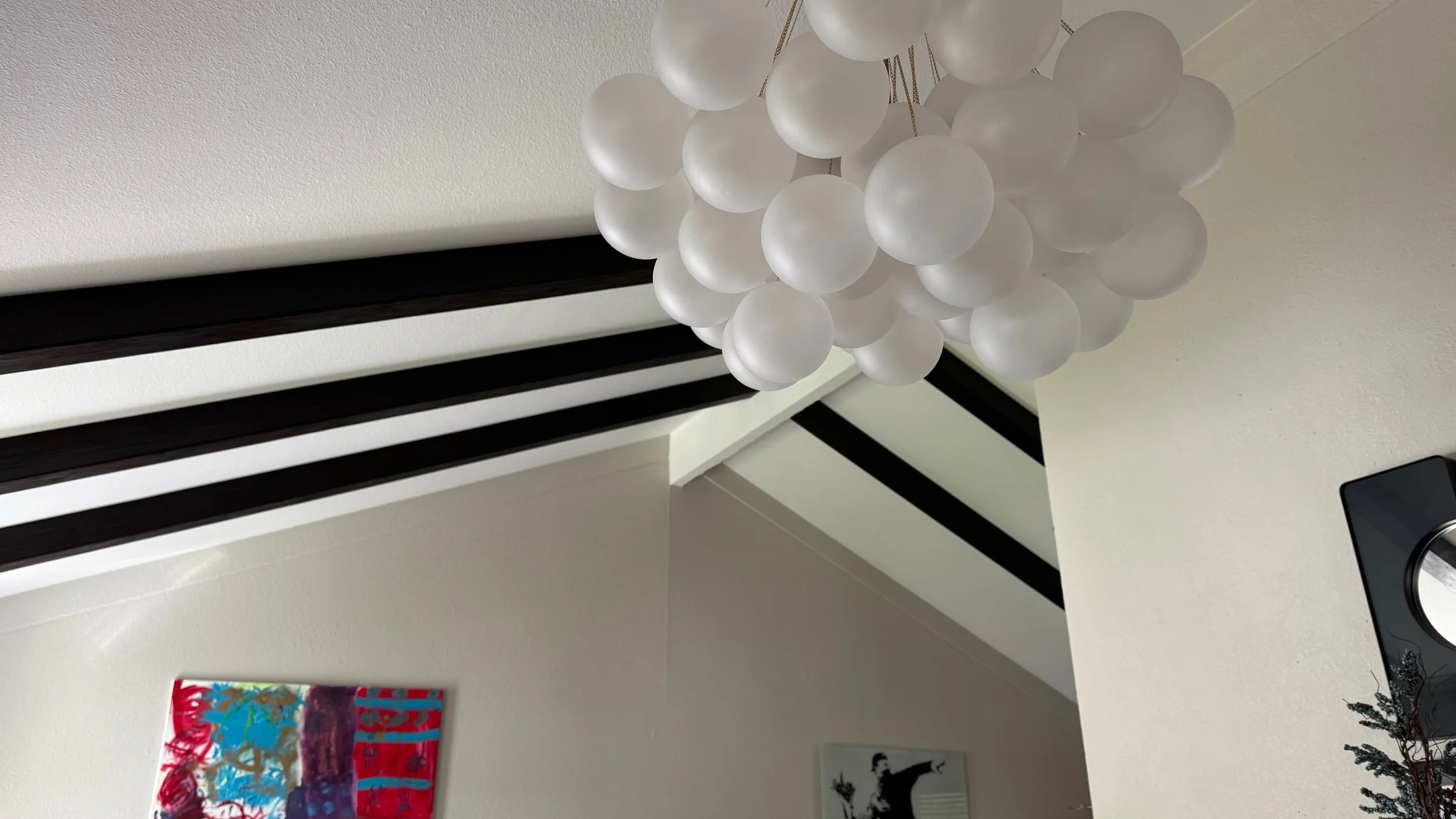 View of a white ceiling with black wooden beams, a modern chandelier with multiple white spherical lights, two colorful abstract paintings on the wall, and part of a table with a speaker and decorative plant.
