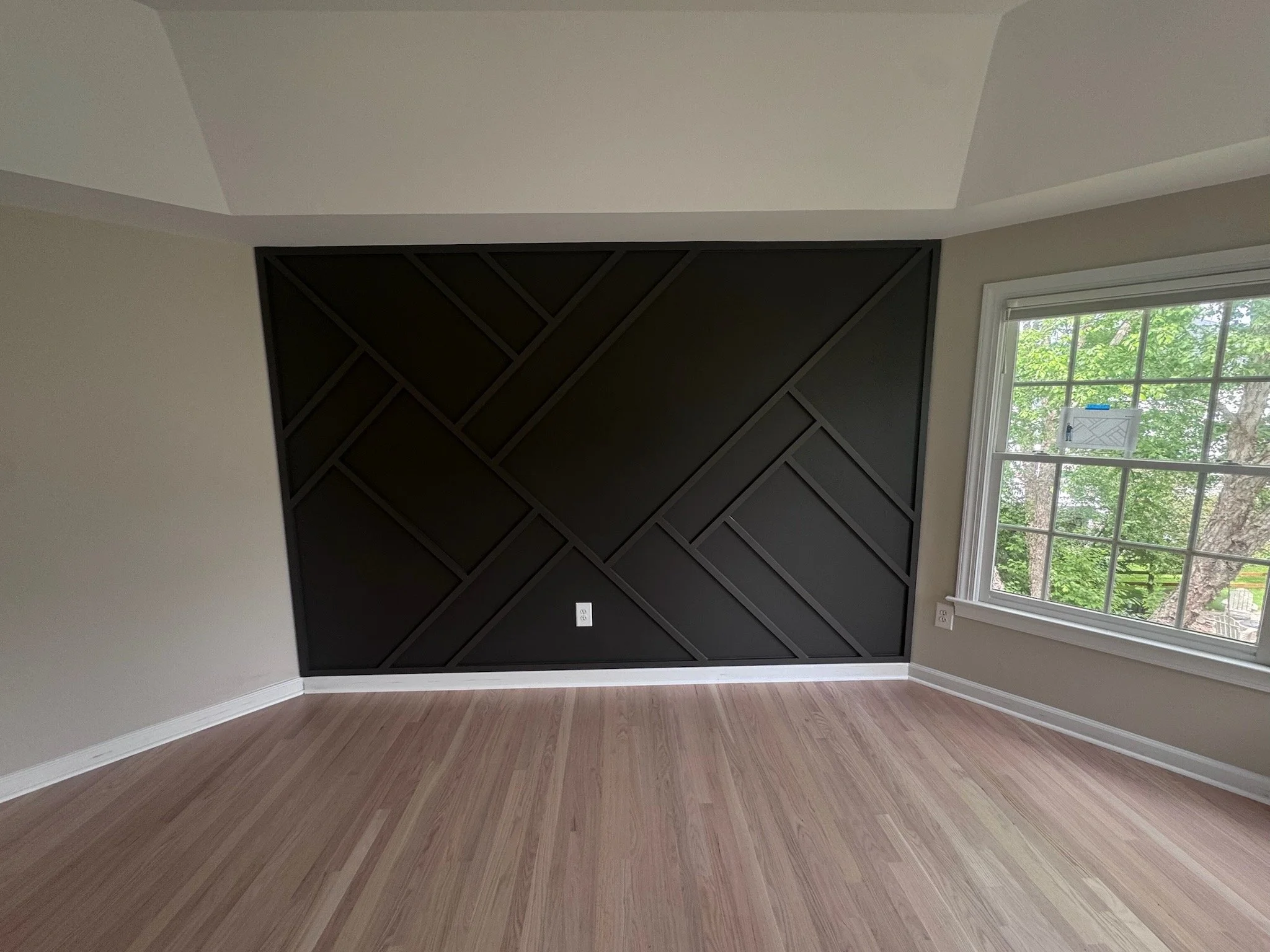 Empty room with a black geometric accent wall, light wooden floor, beige walls, and a large window with a view of green trees outside.