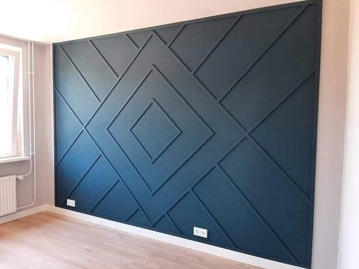 Interior of a room with a dark blue geometric accent wall and a white baseboard, light-colored wooden floor, and a radiator near a window.
