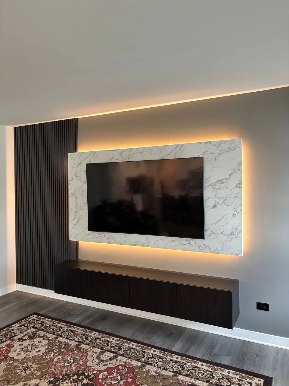 Modern living room with a wall-mounted flat-screen TV surrounded by a white marble frame with backlit lighting, black vertical wall panel on the left, dark floating cabinet beneath, hardwood flooring, and a patterned area rug.