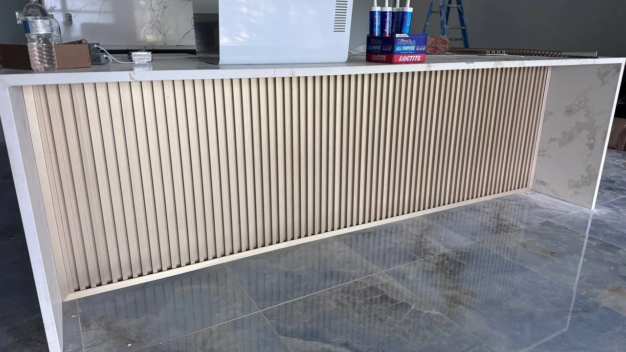 Close-up of a modern kitchen or bar counter with beige vertical wood slats underneath a white countertop, and items like a water bottle, a paper towel roll, milk cartons, and a ladder in the background.