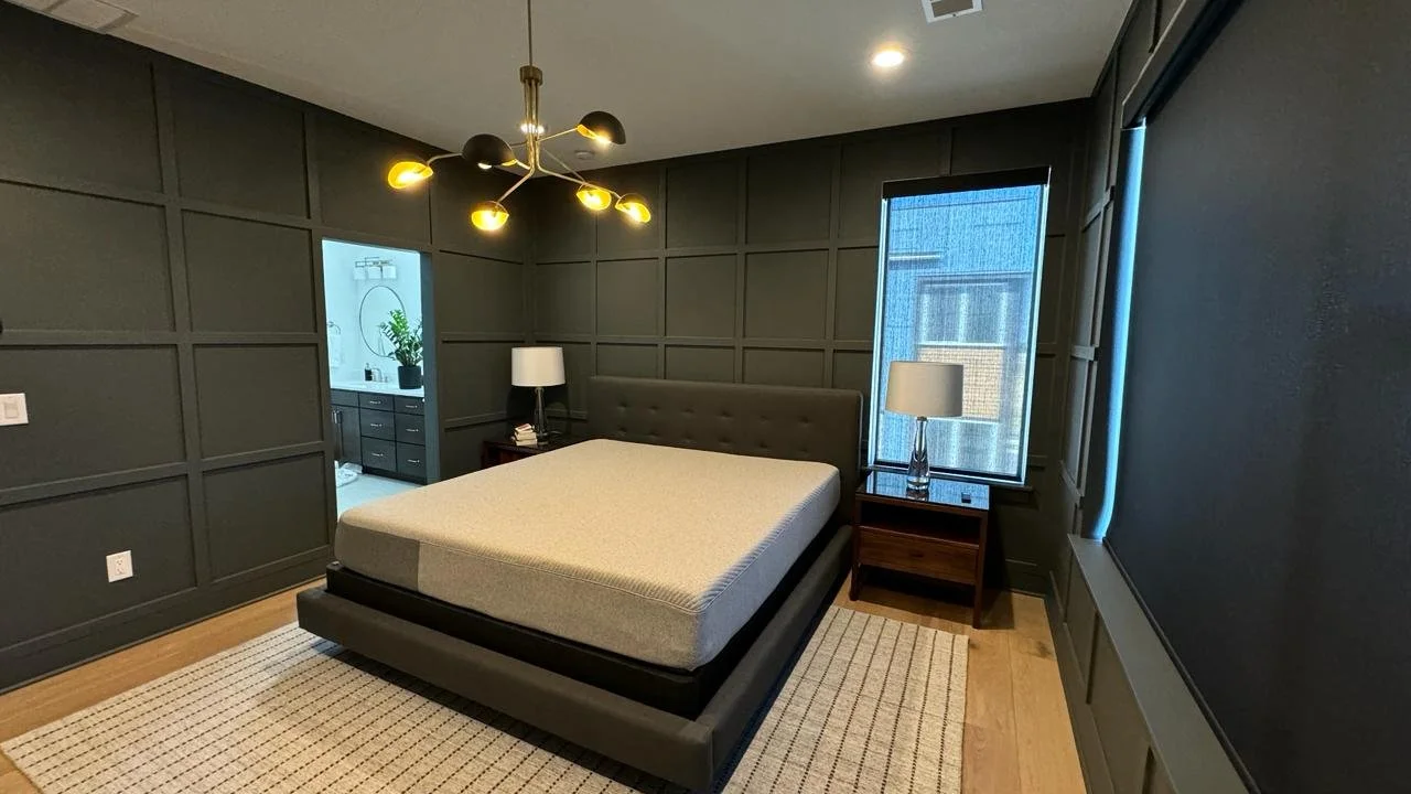 Modern bedroom with dark paneled walls, a bed with beige mattress, and two nightstands with lamps. One window shows a building outside, and a door opens to a bathroom with a mirror and plant. A stylish black and gold chandelier hangs from the ceiling