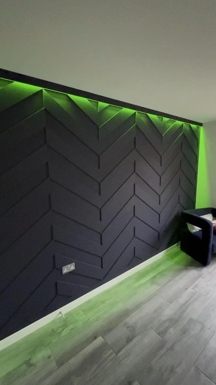 Interior wall with black geometric 3D panels and green LED lighting above and below, next to a small black armchair and a gray wood floor.