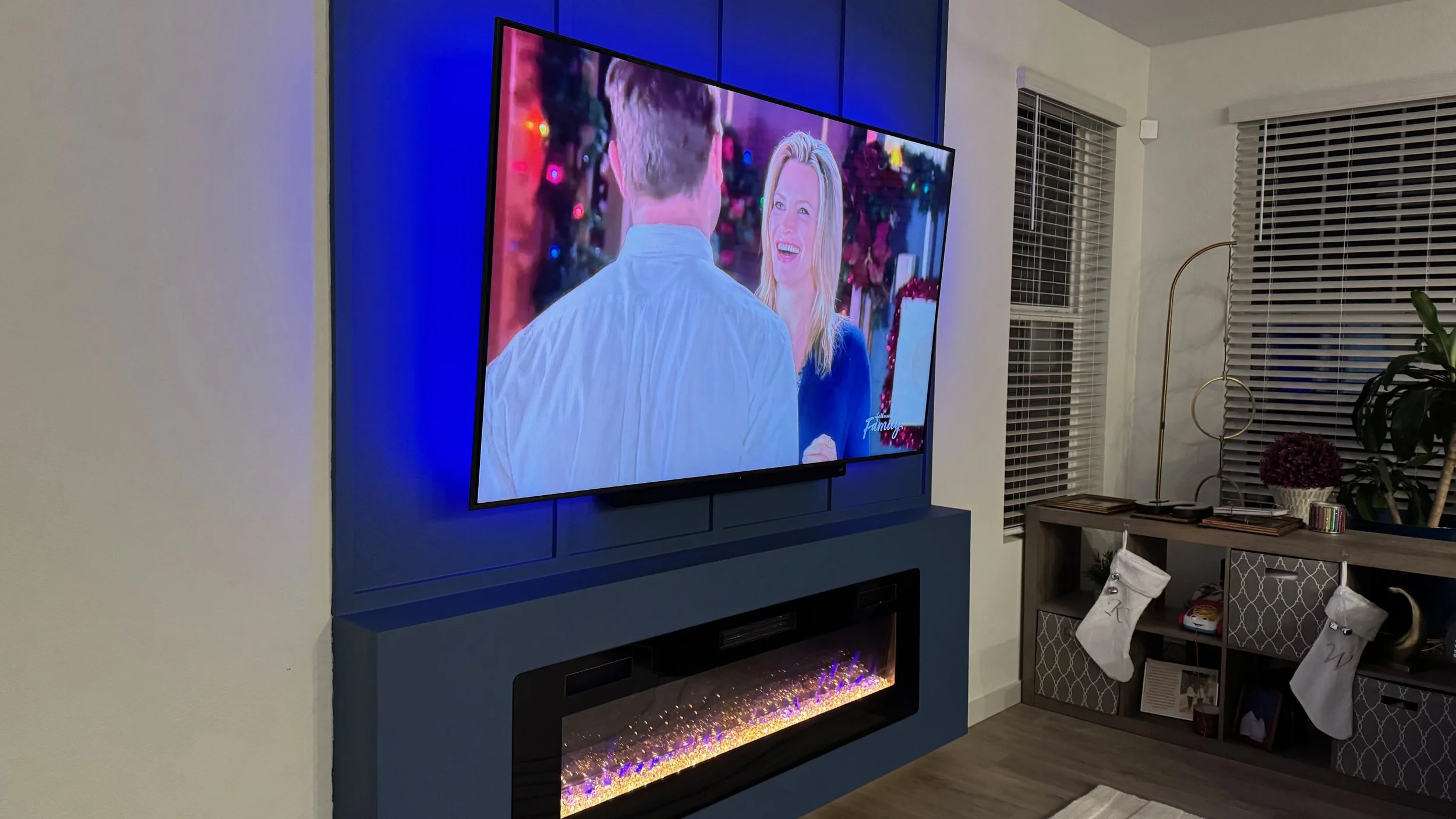 Living room with a large wall-mounted TV showing a scene from a holiday-themed show, decorated with Christmas lights. Below the TV, there is a modern electric fireplace with glowing embers. To the right, there is a wooden sideboard with festive stock