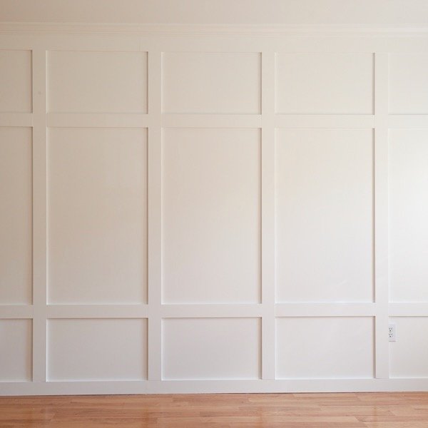 Empty white wall with decorative paneling and a section of baseboard, next to hardwood floor.