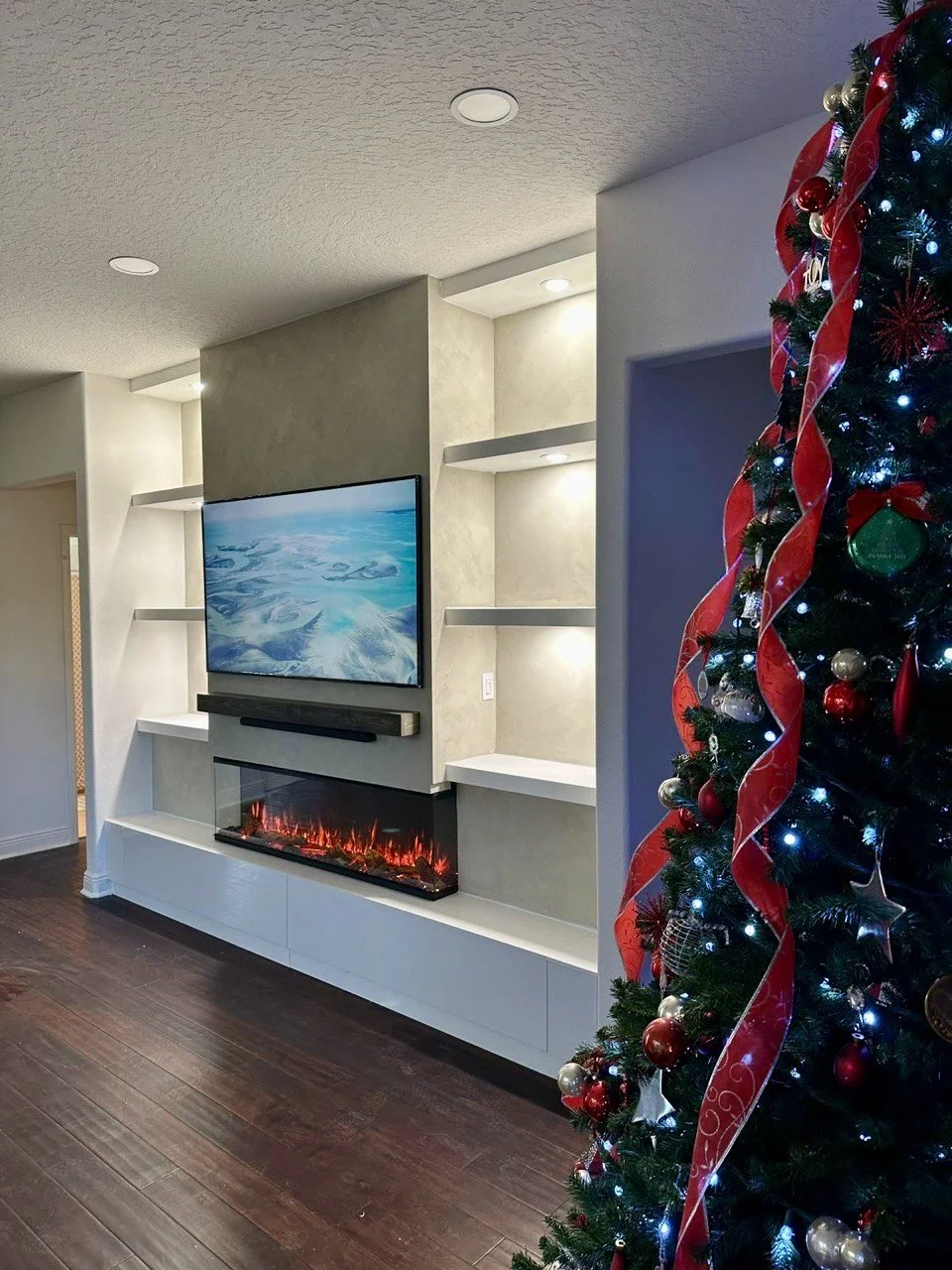 Living room with a Christmas tree decorated with red and gold ornaments, red ribbons, and lights, next to a modern electronic fireplace and a wall-mounted television.