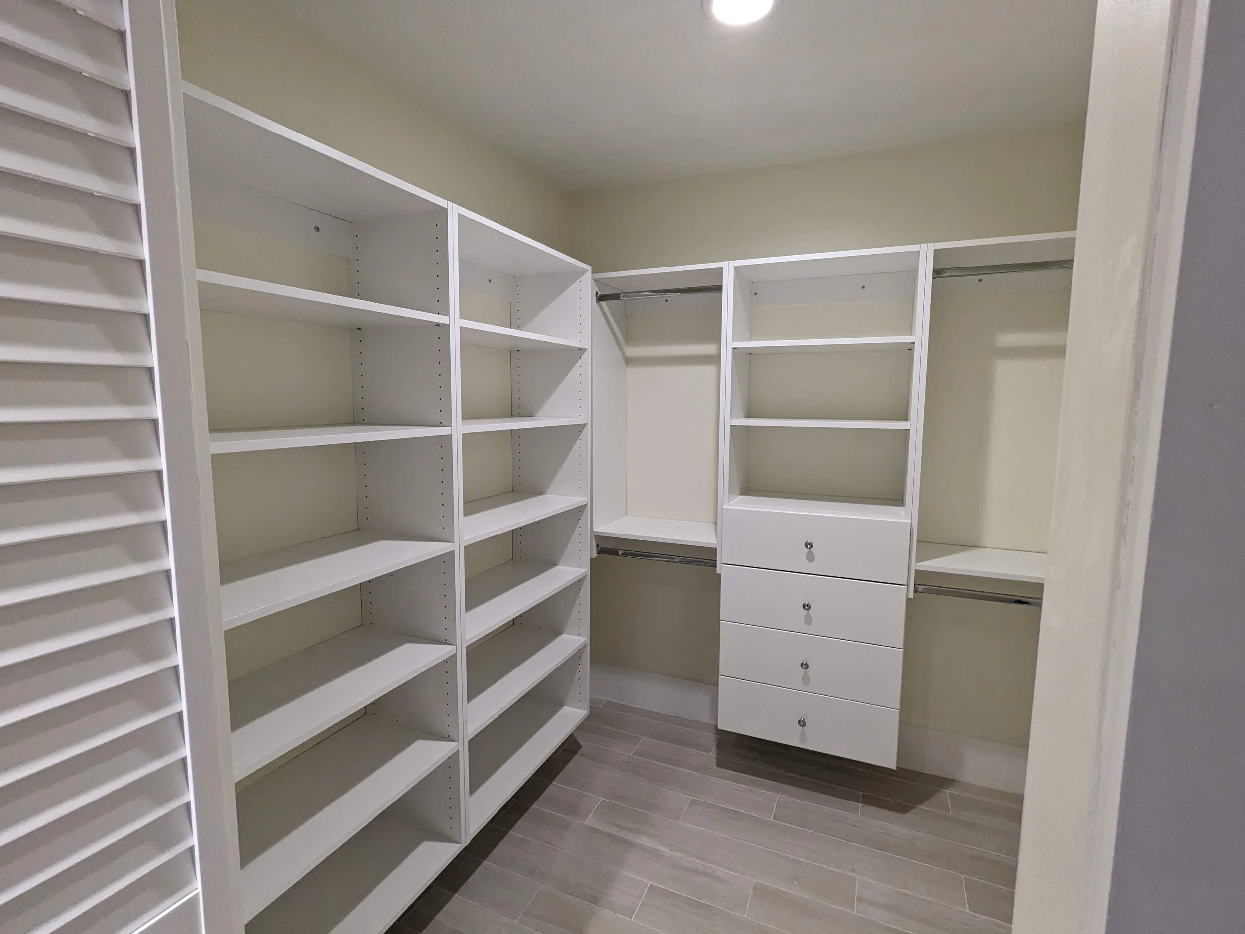 Empty walk-in closet with white built-in shelves, drawers, and hanging rods, with wood-look tile flooring.