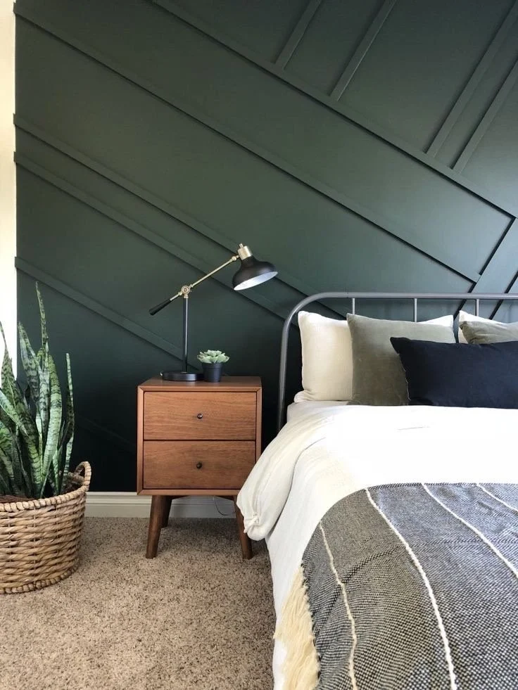A bedroom with a green paneled accent wall, a bed with white bedding and multiple pillows, a small wooden nightstand with a black desk lamp and a potted plant, and a large potted snake plant in a woven basket on a beige carpet.