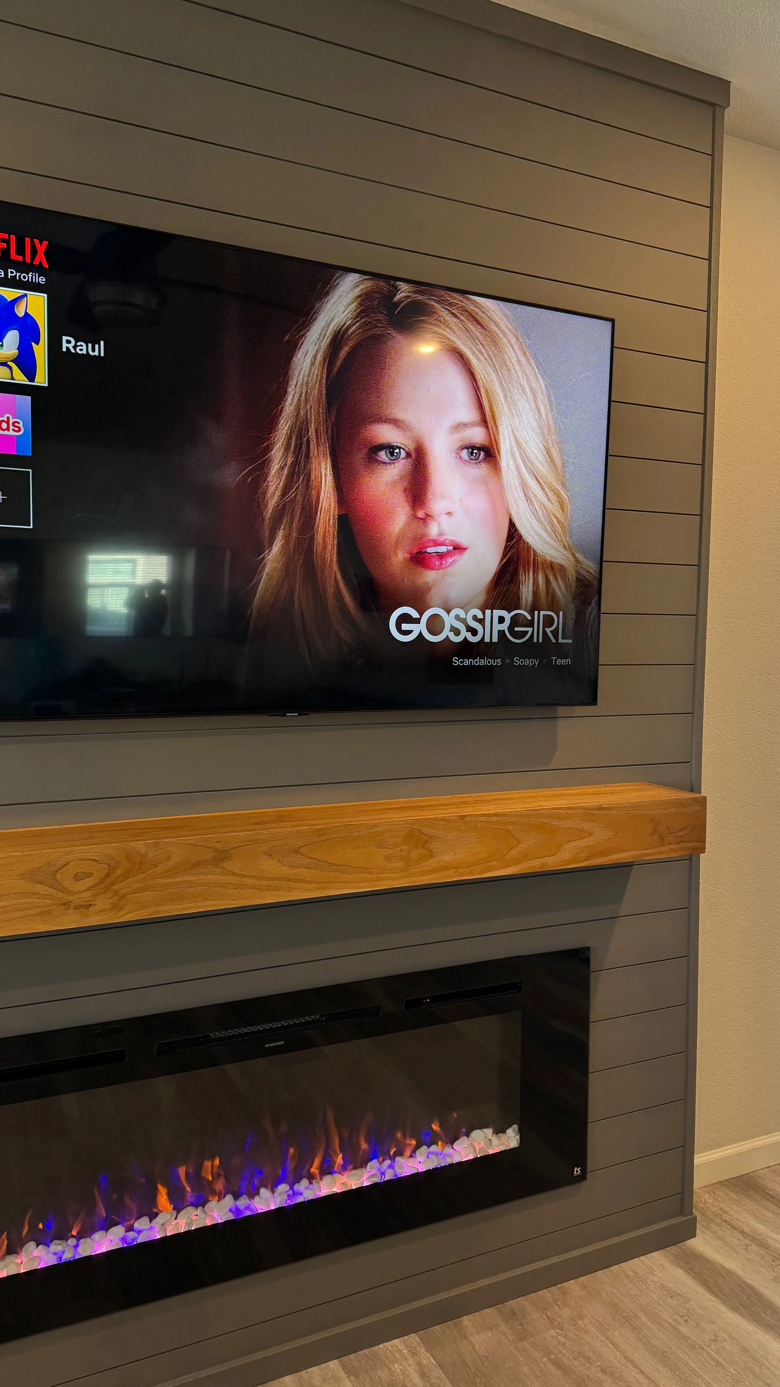 A television displaying the show Gossip Girl with a woman's face on the screen, mounted on a wall above an electric fireplace with colorful flames, and a wooden mantel below the TV.