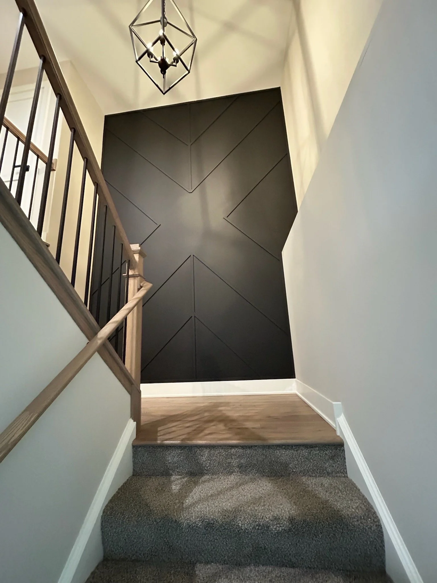 View of a staircase with dark gray carpet, transitioning to a second level with hardwood flooring, black geometric accent wall, white trim, beige walls, and a modern black metal chandelier hanging from the ceiling.