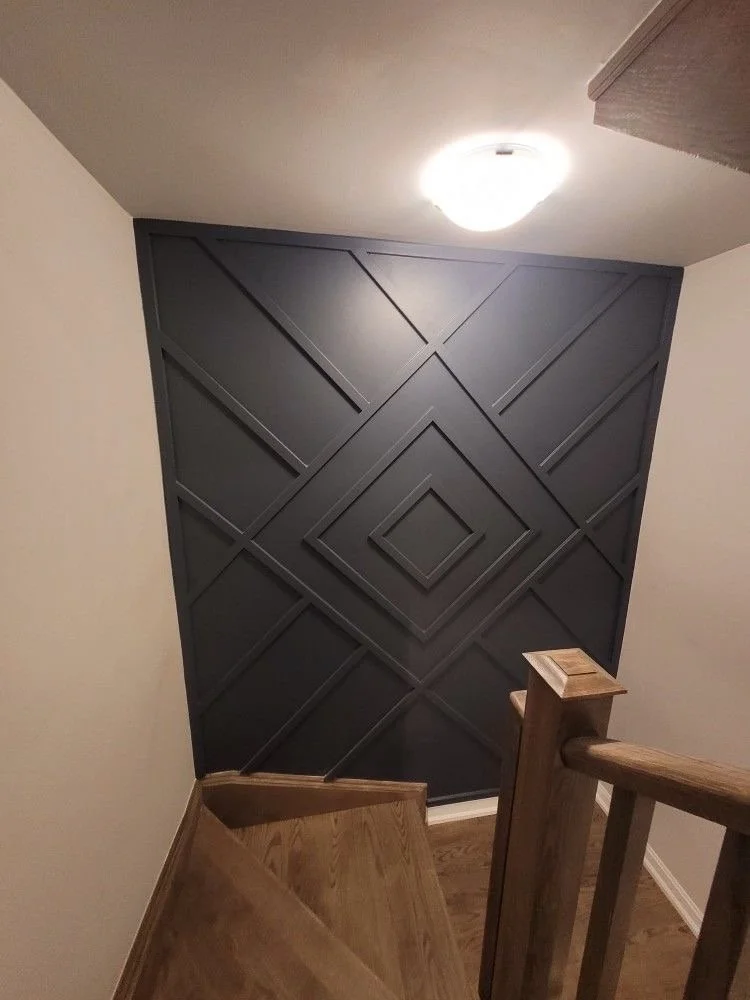 Interior view of a staircase with wooden handrails, leading to a dark accent wall with geometric paneling in a home or apartment.