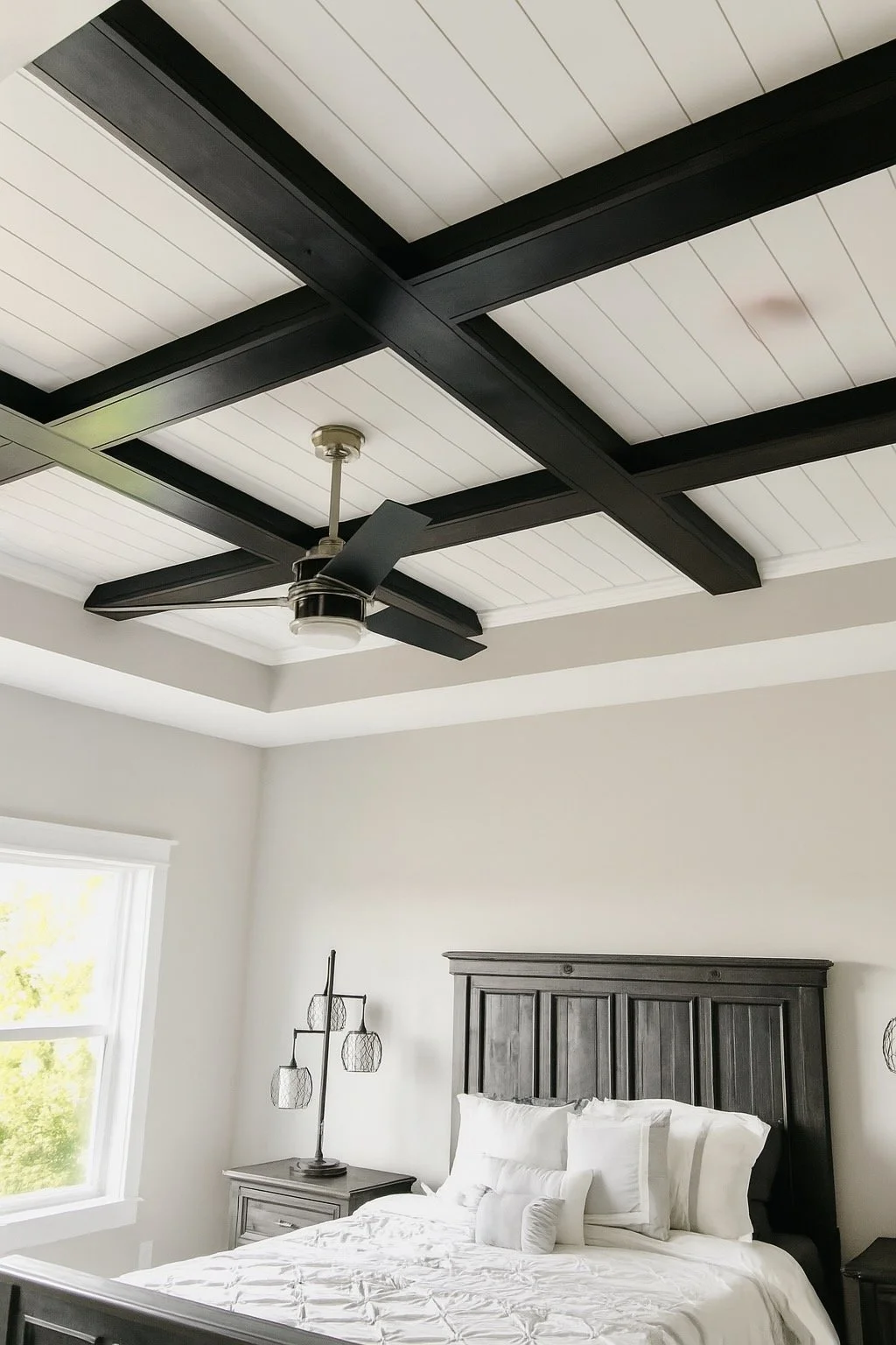 A bedroom with white walls, a black wood-paneled ceiling, a ceiling fan, a bed with white bedding and pillows, a dark gray wooden headboard, and a nightstand with a modern black wire table lamp. A window shows green trees outside.