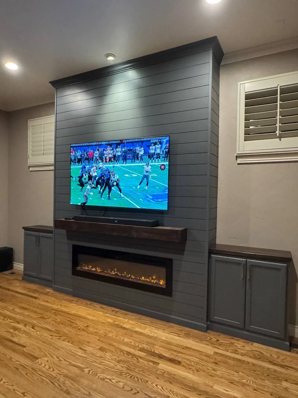 Living room with a mounted television playing a football game on a gray paneled accent wall, below which is a modern electric fireplace, with gray cabinets on either side and hardwood floors.