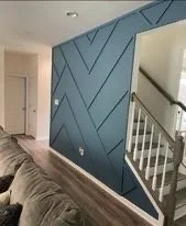 Interior view of a living room with a blue accent wall featuring geometric paneling, a staircase with white railing, and part of a beige sofa.