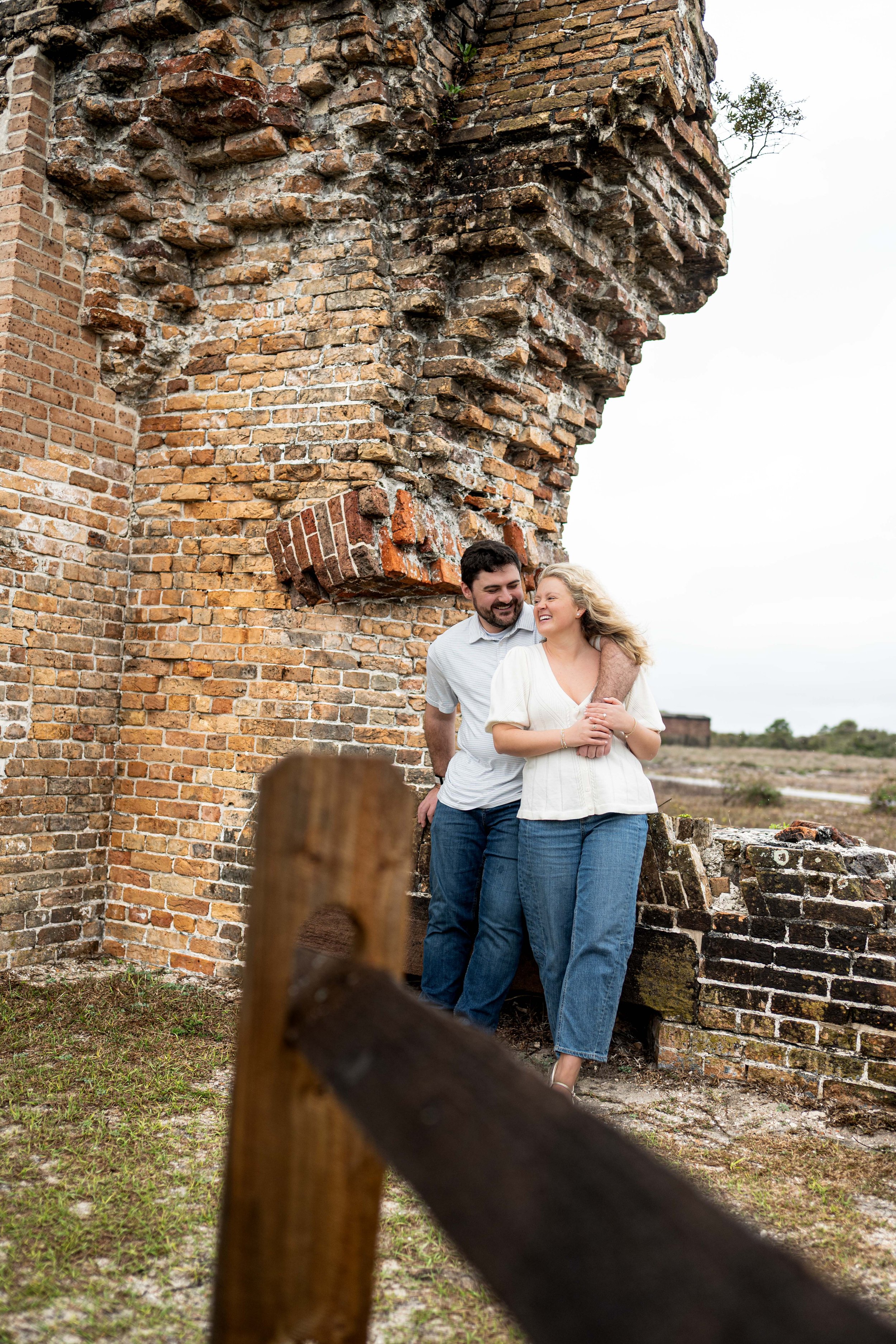 A couple standing close to each other near the ruins of a brick building, smiling and laughing, with an overcast sky in the background.